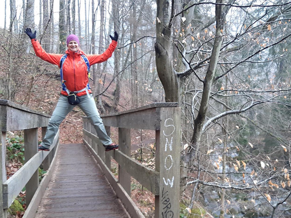 Auf dem Bild stehe ich, Sibylle, mit zum Himmel gereckten Armen auf einer Brücke über ein kleines Tal im Wald. Ich trage Wanderbekleidung mit einer hellen Hose und einer leuchtend roten Jacke.
